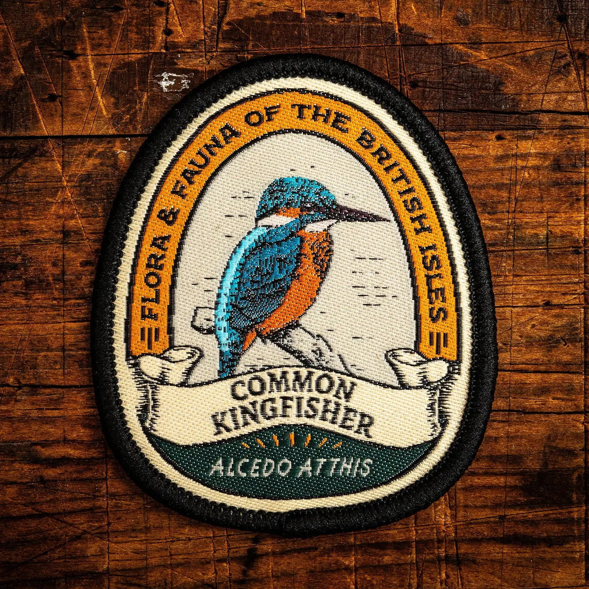 An oval embroidered patch featuring a detailed illustration of a Common Kingfisher on a branch, with the text 'FLORA & FAUNA OF THE BRITISH ISLES' and 'COMMON KINGFISHER' on a rustic dark wood background.