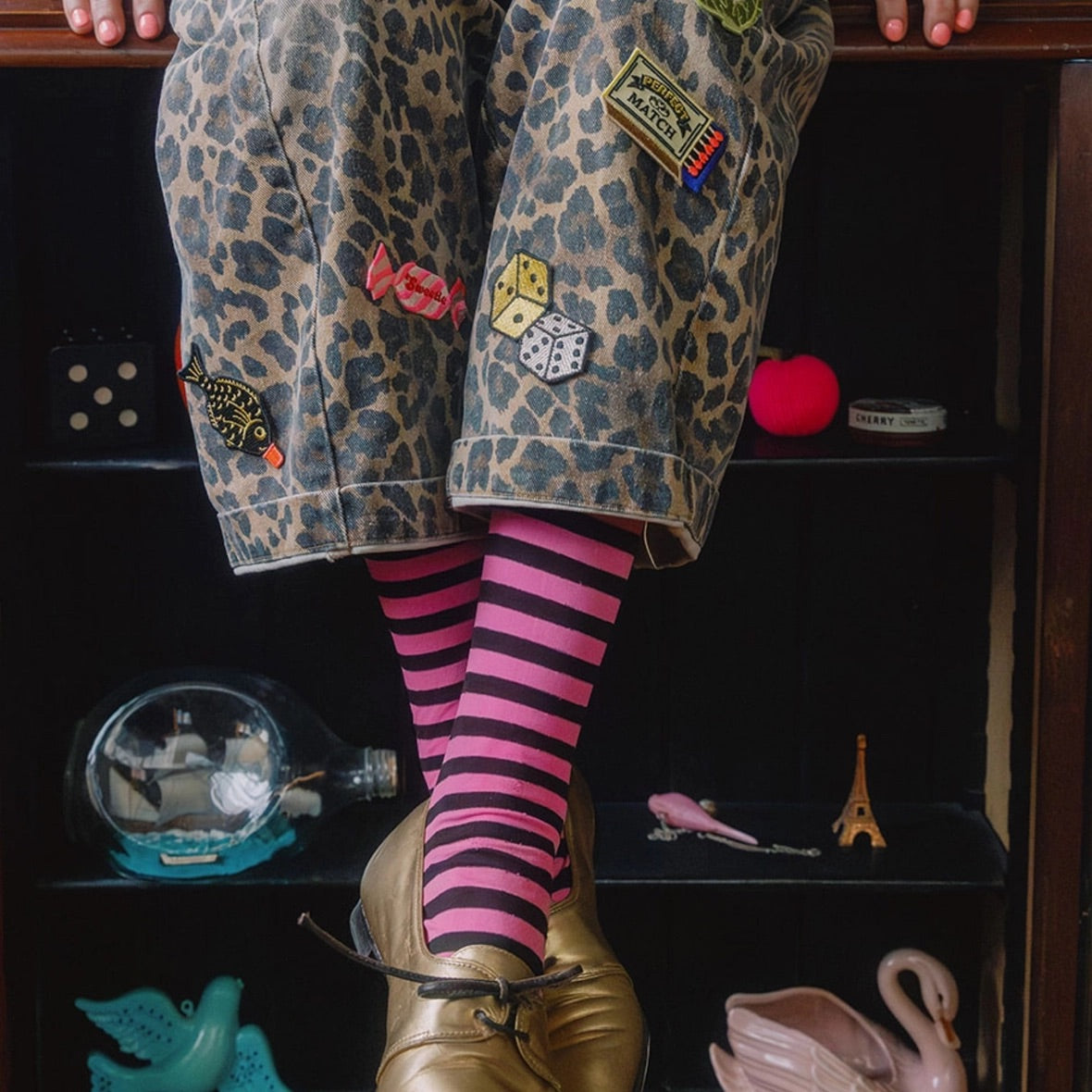A person sitting on a dark shelf wearing leopard-print trousers, bright pink and black striped socks, and shiny gold shoes. The shorts are decorated with several colourful patches, including a fish patch and dice patches.