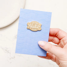 A hand holding a light blue textured card with a gold metal enamel pin that reads 'LET THE Adventure BEGIN'.