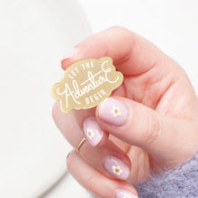 A hand handing a small gold enamel pin that reads 'LET THE Adventure BEGIN' in white script, against a soft white background.