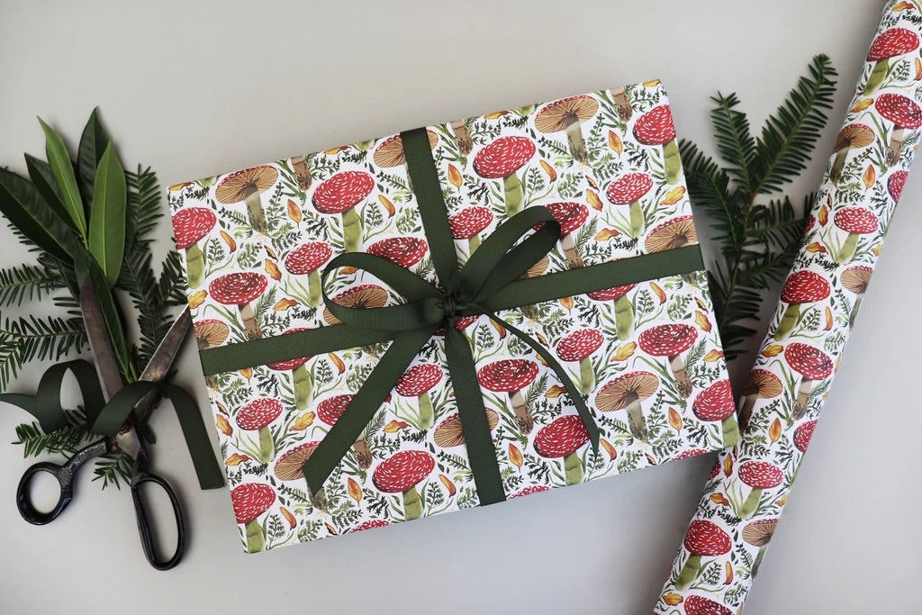 Wrapping paper with a red and green mushroom print, shown wrapped around a gift box tied with a dark green ribbon, alongside a roll of the same paper and black-handled scissors on a light grey background.