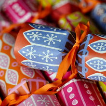 A close-up of a festive paper Christmas cracker with a blue and white snowflake pattern, wrapped with an orange ribbon, nestled among other colourful patterned crackers.