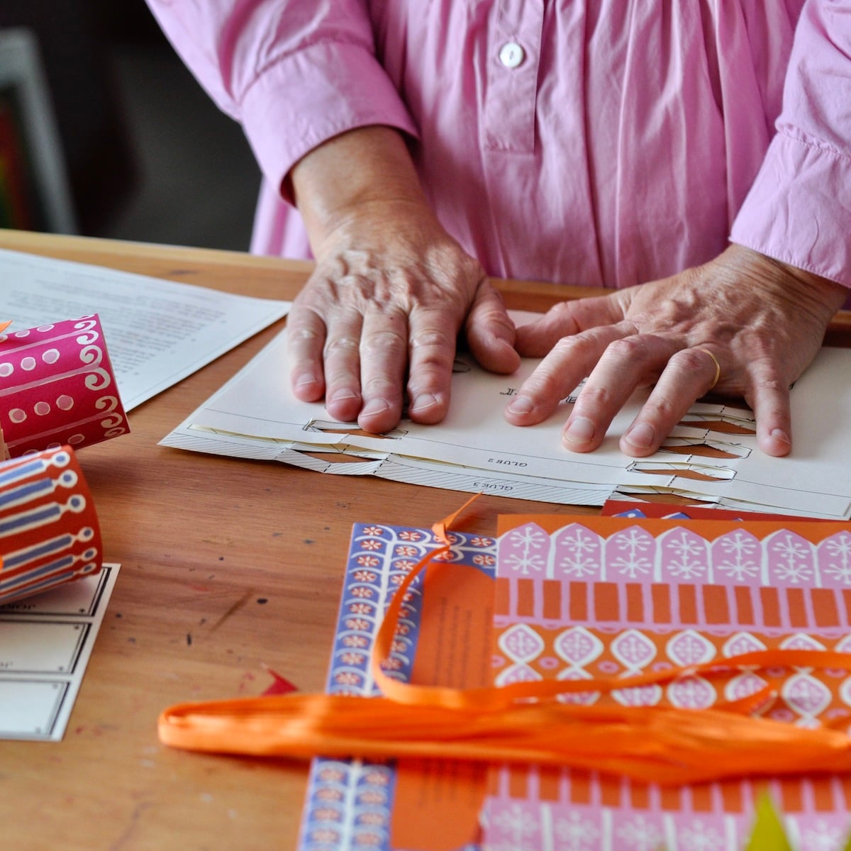 A close-up of a person's hands arranging patterned paper strips on a wooden table, with sheets of patterned paper and orange ribbon in the foreground.
