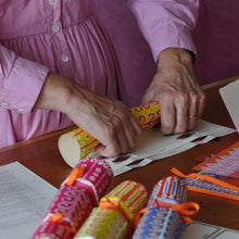 A person is rolling the cracker papers on a wooden table, with several colourful finished crackers and patterned paper strips nearby.