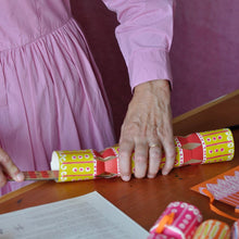 A person is putting the finishing touches to their Christmas cracker on a wooden table, with other wrapped crackers and printed papers nearby.
