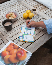 A person's hand writes on a colorful sunflower themed meal  planner or  on a wooden outdoor table, surrounded by lemons, apples, a cup of coffee, and an open cook book.
