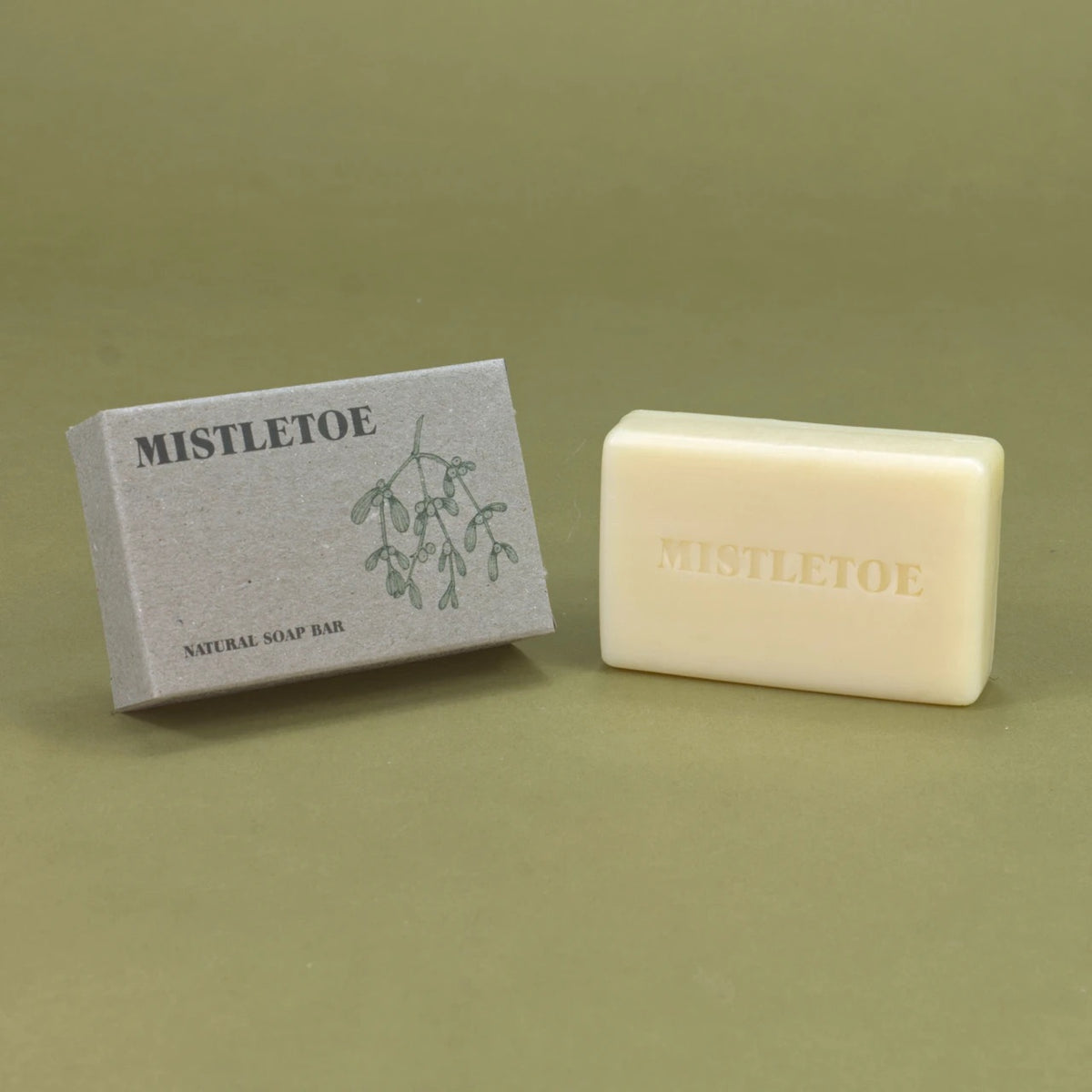 A bar of Mistletoe natural soap bar in an off-white cream color, shown next to its textured gray packaging box with a green mistletoe illustration, set against a solid olive green background.