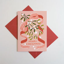 A pink Christmas card with a red striped ribbon and green mistletoe illustration, placed on top of a red envelope against a light gray background.