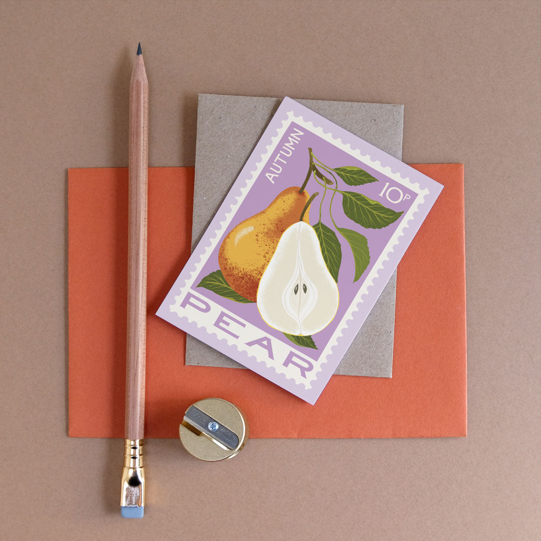 A flat-lay of a pear-themed card on top of a kraft envelope and orange cardstock, with a wooden pencil and a small sharpener arranged around it on a taupe background.