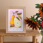 A framed art print featuring an autumn pear illustration in a light wood frame, styled on a wooden table next to sunflowers against a beige wall.