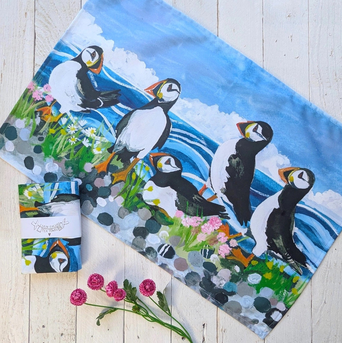 Tea towel printed with a colourful painting of puffins on a rocky shore with blue ocean waves. The fabric is laid flat on a white wooden surface next to a packaged tea towel with a white paper band and pink flowers.