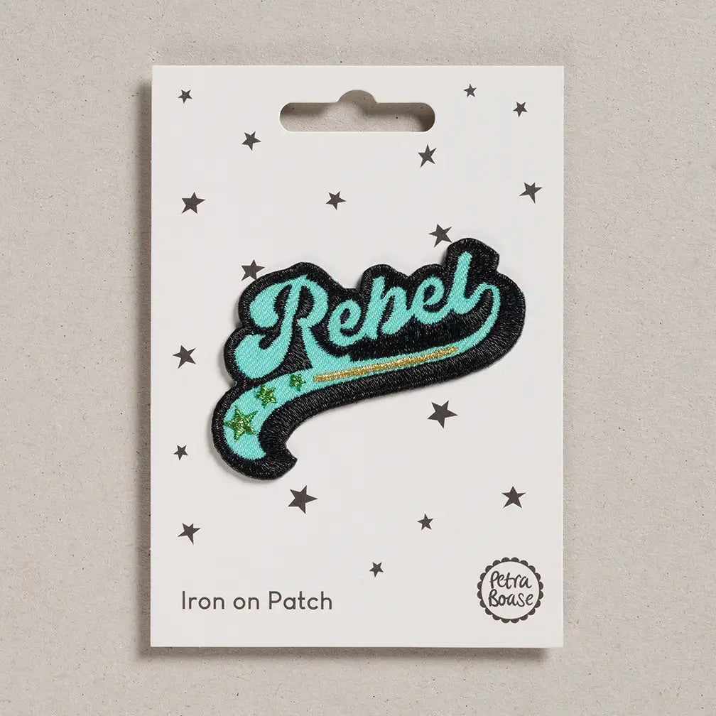 A 'Rebel' iron-on patch by Petra Boase, featuring a turquoise and black script design with a gold glitter stripe and green stars, displayed on its white backing card.