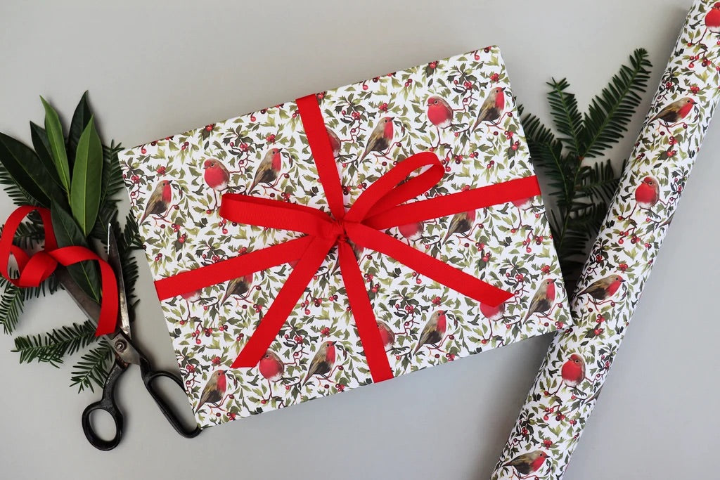 A gift-wrapped box and a roll of wrapping paper on a light gray background, both printed with a white base and a repeating pattern of red robins, green leaves, and small ornaments. A red ribbon is tied around the box, and holiday greenery and scissors are placed around it.