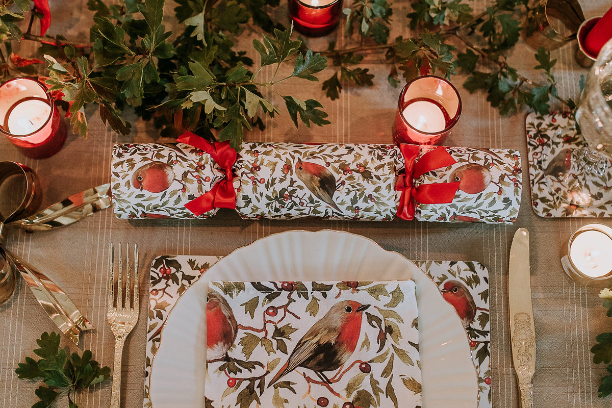 A festive Christmas table setting featuring a Christmas cracker with red robin design, tied with red ribbons. Matching place settings with white plates, gold cutlery, and a red robin printed napkin are arranged on a rustic wooden table decorated with green foliage and red candles.