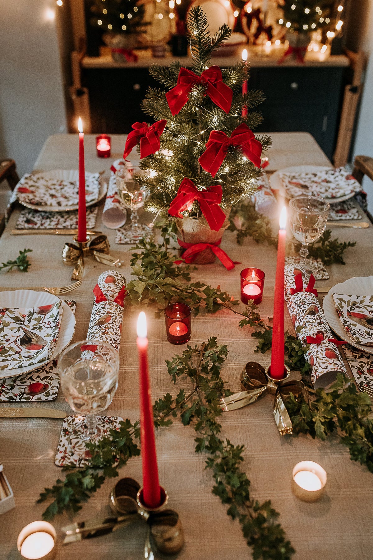 A festive Christmas table setting with a small decorated evergreen tree centerpiece, red candles, green garlands, patterned plates, and red-and-white napkins in a warmly lit dining room.