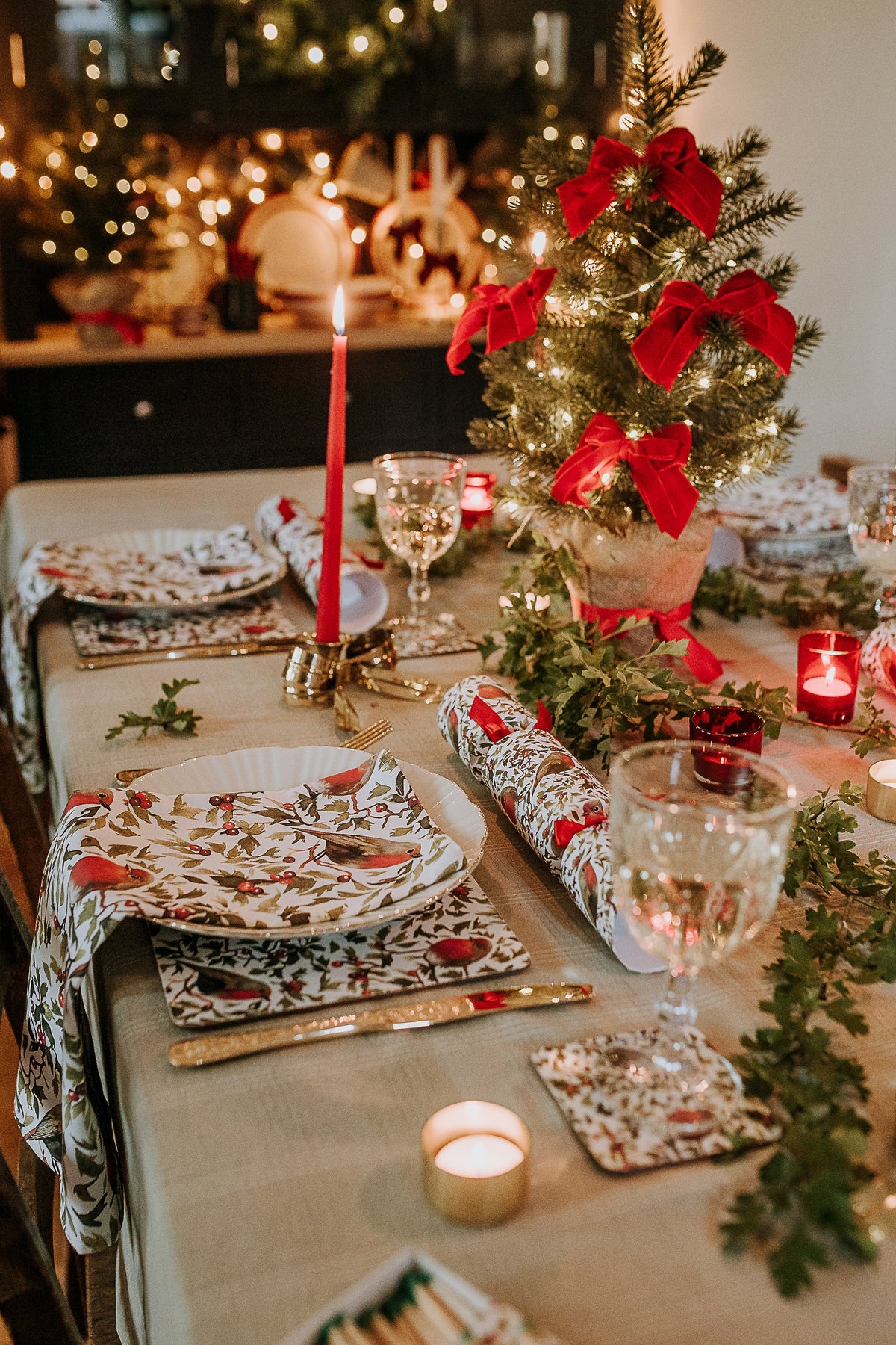 A festive Christmas table setting with matching patterned napkins, Christmas crackers, decorated with a small lit tree, red bows, candles, and greenery in a warmly lit indoor setting.