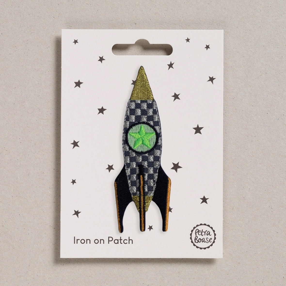 A rocket-shaped iron-on patch displayed on its white backing card, which features a star pattern and the text 'Iron on Patch' and 'Petra Boase'. The patch has a black and grey checkered body, a green star centre, and gold and black accents.