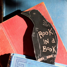 Black leather bookmark in the shape of a rook with the text 'rook in a book' poking out of an old book and set against a background of vintage books. 