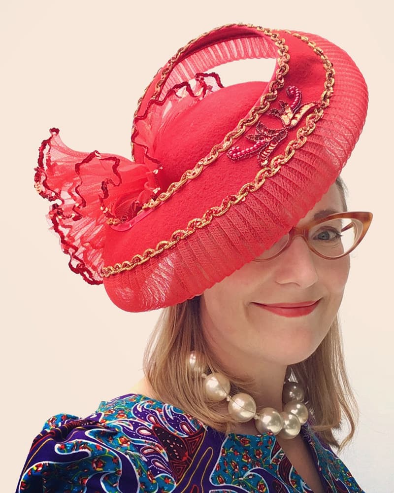 Illustrator Sarah McIntyre wearing a bright red fascinator hat with gold chain trim and a matching red ruffled bow, paired with a colourful paisley blouse and pearl necklace against a plain light background.