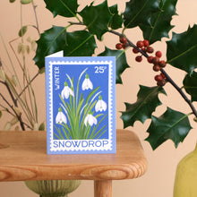 Mini card featuring snowdrop vintage stamp style design in blue, shown on wooden bench with holly in the background.