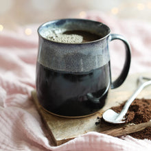 Black mug with white decoration around the top with coffee on a wooden coaster with a spoon and coffee grounds.