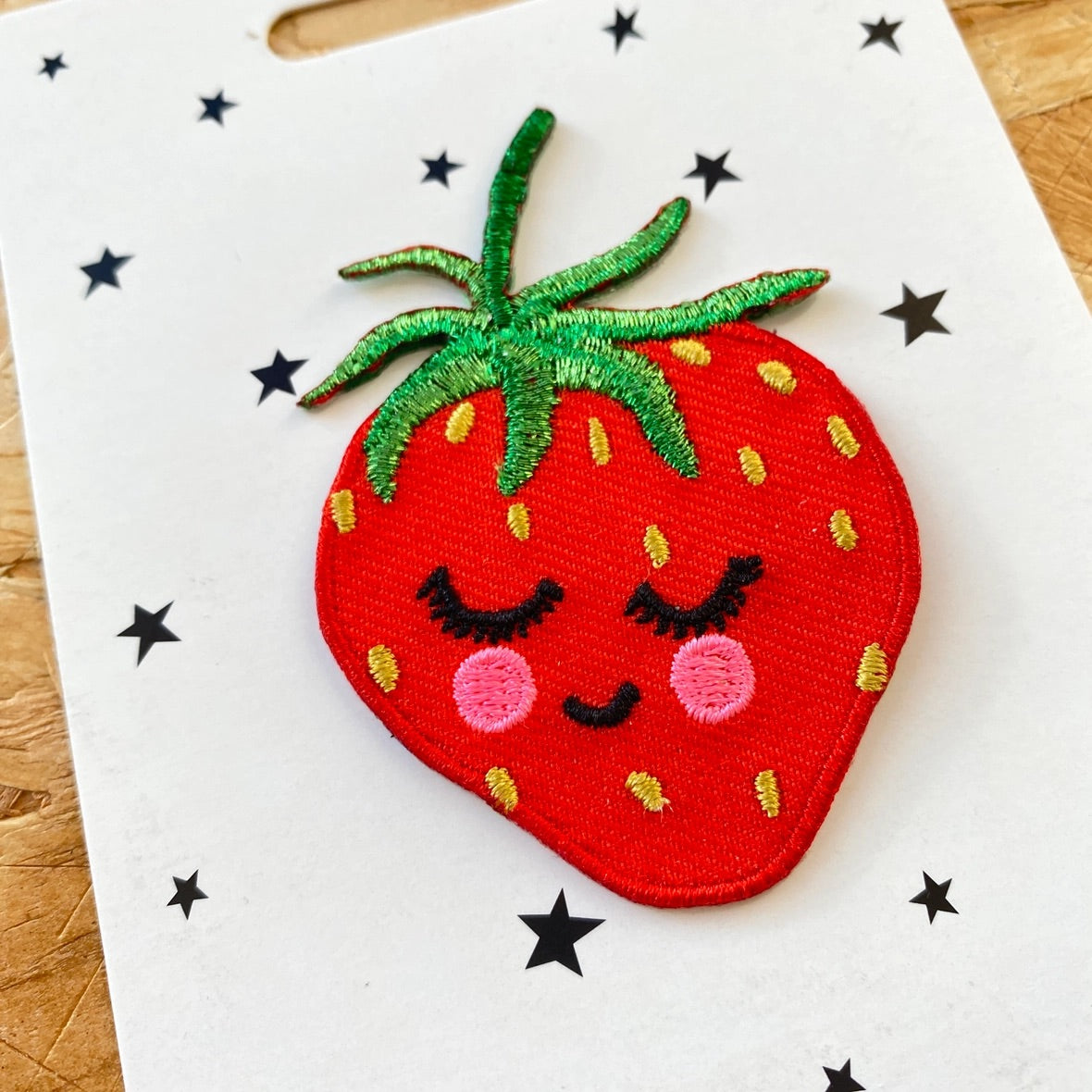 A red embroidered strawberry patch with a smiling face, pink cheeks, and green leafy top, displayed on a white card with black star cutouts over a wooden surface.