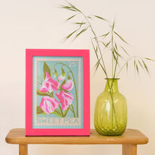 A framed art print featuring pink sweet pea flowers on a light blue background, styled on a wooden table next to a green glass vase with dried grass against a cream wall.