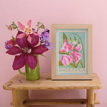 A framed vintage-style sweet pea art print sits on a wooden table next to a green mug filled with flowers against a solid pink wall.