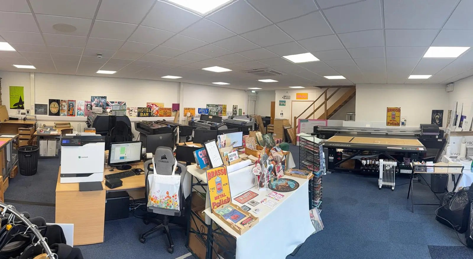 A wide interior view of a large office or workshop space with desks, chairs, computers, a staircase, and a long printing or cutting machine. A small display table in the foreground holds colorful printed materials and books.