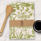 Packaged tea towel with green floral design and text, wooden spoon, and cup of coffee on a light background