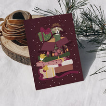 A Christmas card featuring a dog peeking out from a stack of wrapped presents, styled on a white textured surface with a wood slice and pine branches behind.