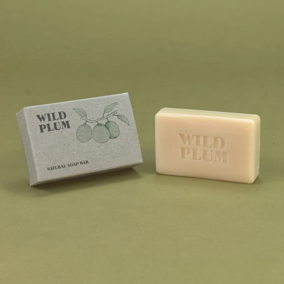 A rectangular bar of natural soap in a pale peach-beige color next to its textured gray packaging box. The box features a green illustration of plums and the embossed text 'WILD PLUM'.