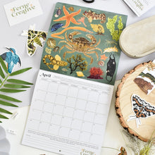 A printed monthly calendar page with a sea-creature illustration cover and visible April grid, styled on a light surface with decorative stickers, leaves, and wood slices. The brand name Ferne Creative appears on the calendar and nearby card.
