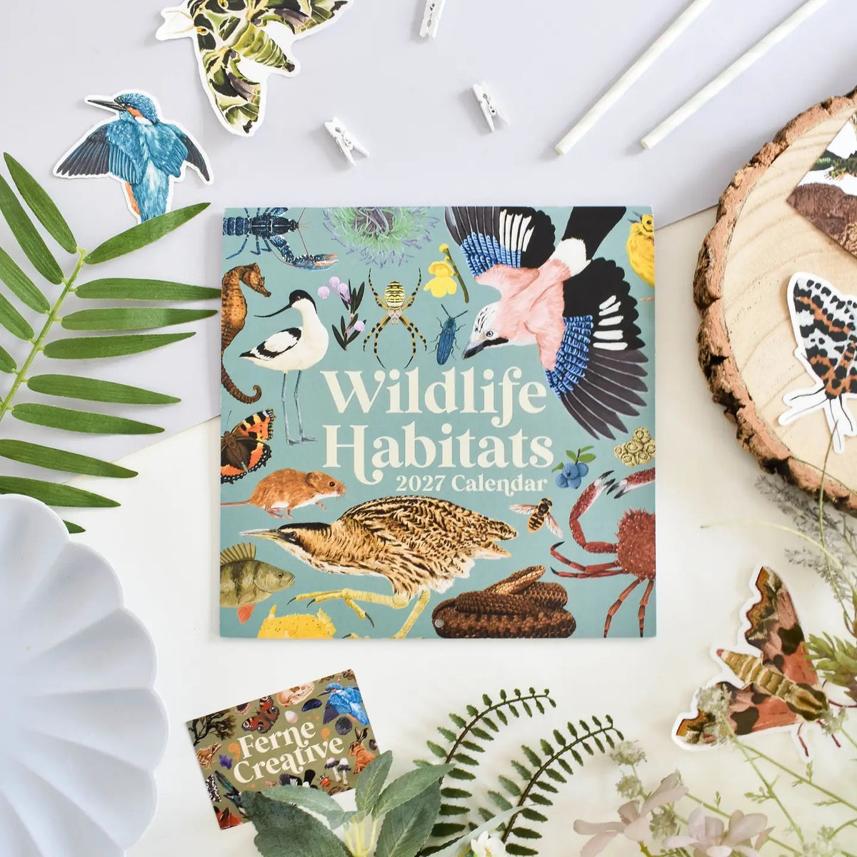 A square 2027 Wildlife Habitats calendar lies on a white surface surrounded by decorative natural props, including leaves, a wood slice, paper clips, and butterfly and bird stickers. 