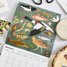 A January calendar page featuring a detailed illustration of various birds and fish on a muted green background, styled on a light surface with partial props around it.