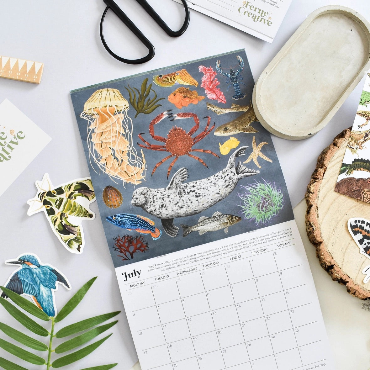 An illustrated July calendar page featuring ocean and sea life illustrations, styled in a flat lay with scissors, a small tray, paper cutouts, and natural props on a light surface.