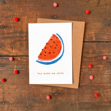 Greeting card with watermelon design and 'You make me smile' text on a wooden surface with decorative items.