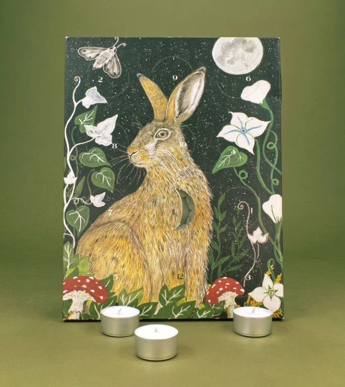 Advent calendar featuring a painted hare in a woodland scene with a moon, flowers, leaves, and red-and-white mushrooms, displayed against an olive green background with three silver tea light candles in front.