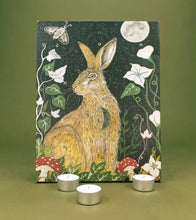 Advent calendar featuring a painted hare in a woodland scene with a moon, flowers, leaves, and red-and-white mushrooms, displayed against an olive green background with three silver tea light candles in front.