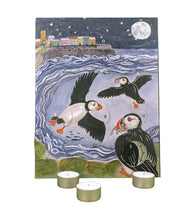 Advent calendar featuring a painted puffin scene at night, with three small tealight candles placed in front on a white background.