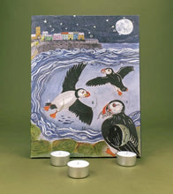 Advent calendar featuring puffins over stylized blue water with a coastal town and moon in the background, displayed on an olive green surface with three small silver tea lights in front.