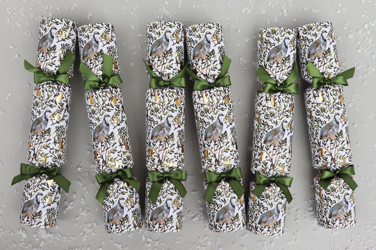 Six packaged Christmas crackers arranged  on a light grey textured background. Each cracker is wrapped in white paper with a partridge in a pear tree print and tied with an olive green ribbon.