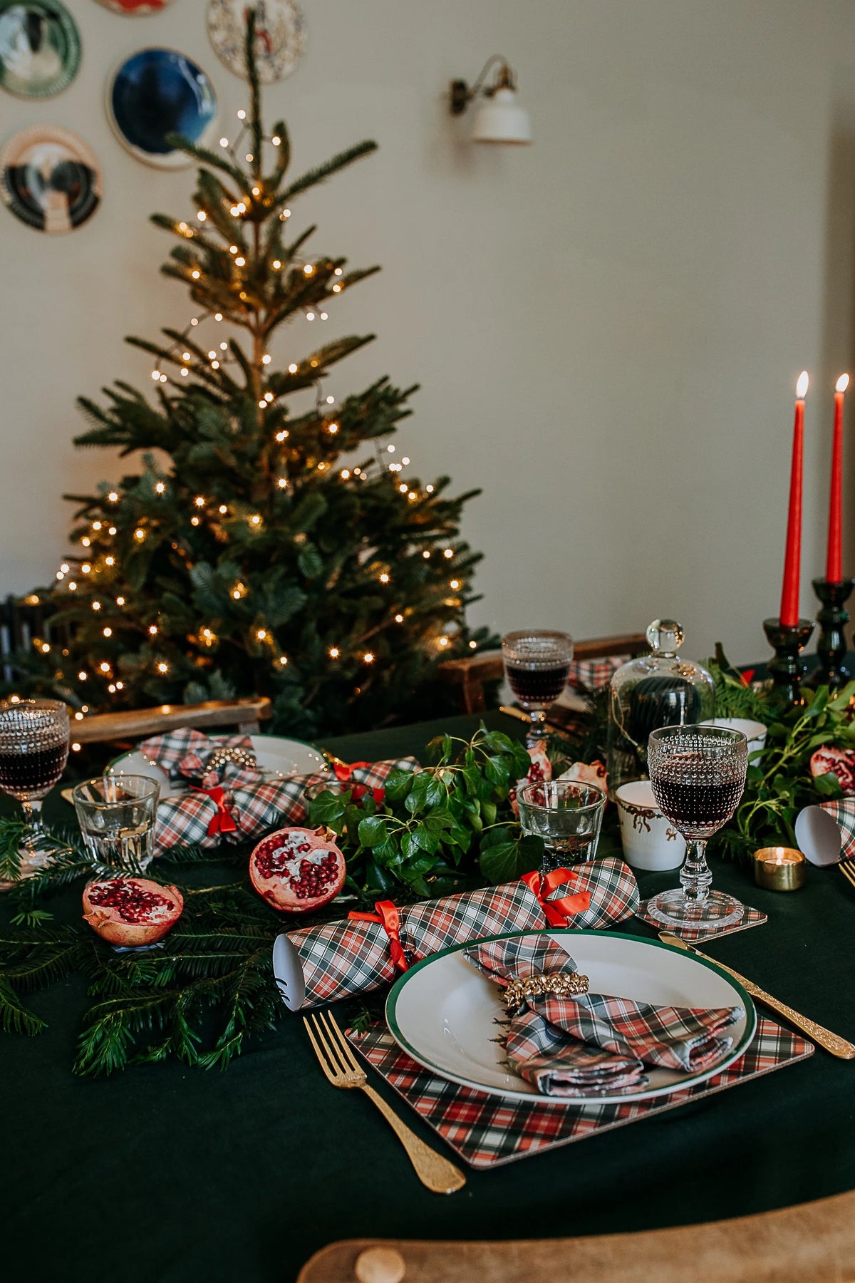 Christmas table with Christmas tree with lights in the background. Red and green tartan Christmas crackers with red bow details. 